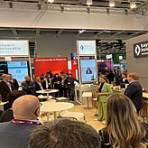 Auf dem Bild ist die Rückansicht von Personen zu sehen, welche sich vor einem Messestand einen Vortrag anhören.