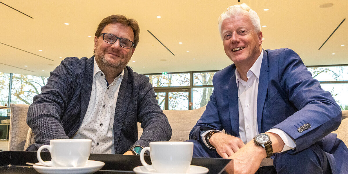 Cluster-Leiter Holger Czuday (links) und Cluster-Sprecher Thomas Regnet (rechts) sitzen an einem Tisch mit Kaffeetassen, blicken freundlich in die Kamera und lächeln