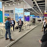 Auf dem Bild sind ein paar Personen auf einem Messegelände zu sehen, welche sich eine Ansprache anhören.