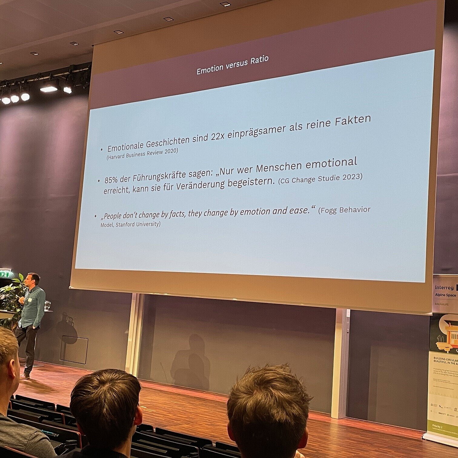 Aufnahme einer Konferenz mit Sprecher auf der Bühne, hinter ihm die Leinwand mit Präsentation und ein Roll-up. 