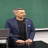  Ein Mann im Anzug steht vor einem schwarzen Brett und präsentiert Informationen.