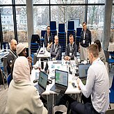 Auf dem Bild ist eine Gruppe von Menschen zu sehen, welche an einem Tisch vor ihren Laptops sitzen und arbeiten.