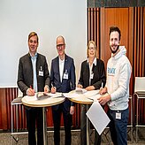 Vier Personen stehen um einen Tisch, auf dem sich Dokumente und Stifte befinden. Im Hintergrund sind eine weiße Tafel oder Leinwand sowie Holzverkleidung zu sehen.