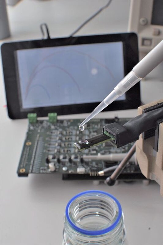 Nahaufnahme eines Tropfens an der Spitze einer Pipette vor einer Platine mit elektronischen Bauteilen und einem Bildschirm im Hintergrund