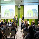 Auf dem Bild ist ein Veranstaltungsraum mit Reihenbestuhlung und zwei Leinwänden zu sehen. Vorne stehen drei Personen mit Mikrofon, welche einen Vortrag halten.