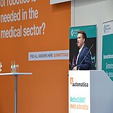 Eine Person steht an einem Rednerpult und hält eine Präsentation. Im Hintergrund sind verschiedene Texte und Grafiken zu sehen