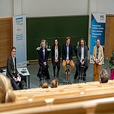 Fünf sitzende Personen auf Hockern und eine stehende Person in einem Hörsaal mit zwei Roll-ups im Hintergrund