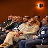 Gruppe von Personen sitzt in einem Raum mit oranger Wand und rundem Wandlicht, einige tragen Namensschilder und eine Person hält ein Smartphone in der Hand