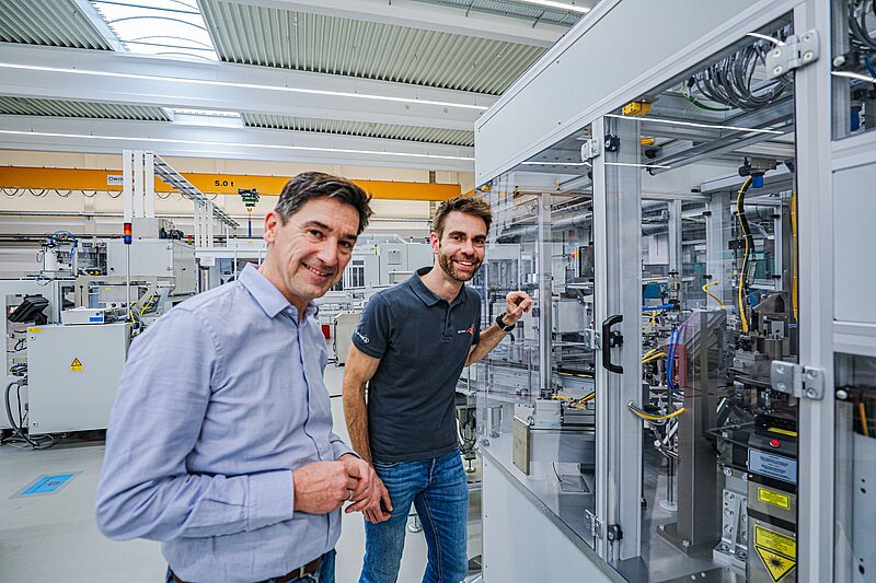 Zwei Männer stehen in einem hellen Industrie- oder Laborraum vor einer komplexen technischen Anlage mit Kabeln und Steuergeräten