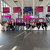 Auf dem Bild ist eine Gruppe von Personen zu sehen, welche auf einer Anhöhe in einem Veranstaltungsgebäude sitzen. Hinter Ihnen hängen verschiedene Monitore und Werbebanner an der Decke.