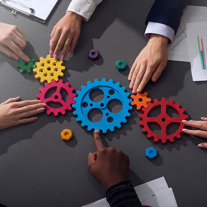 Aus der Vogelperspektive sieht man einen Tisch auf dem bunte Zanhräder liegen die von verschiedenen Händen berührt werden.