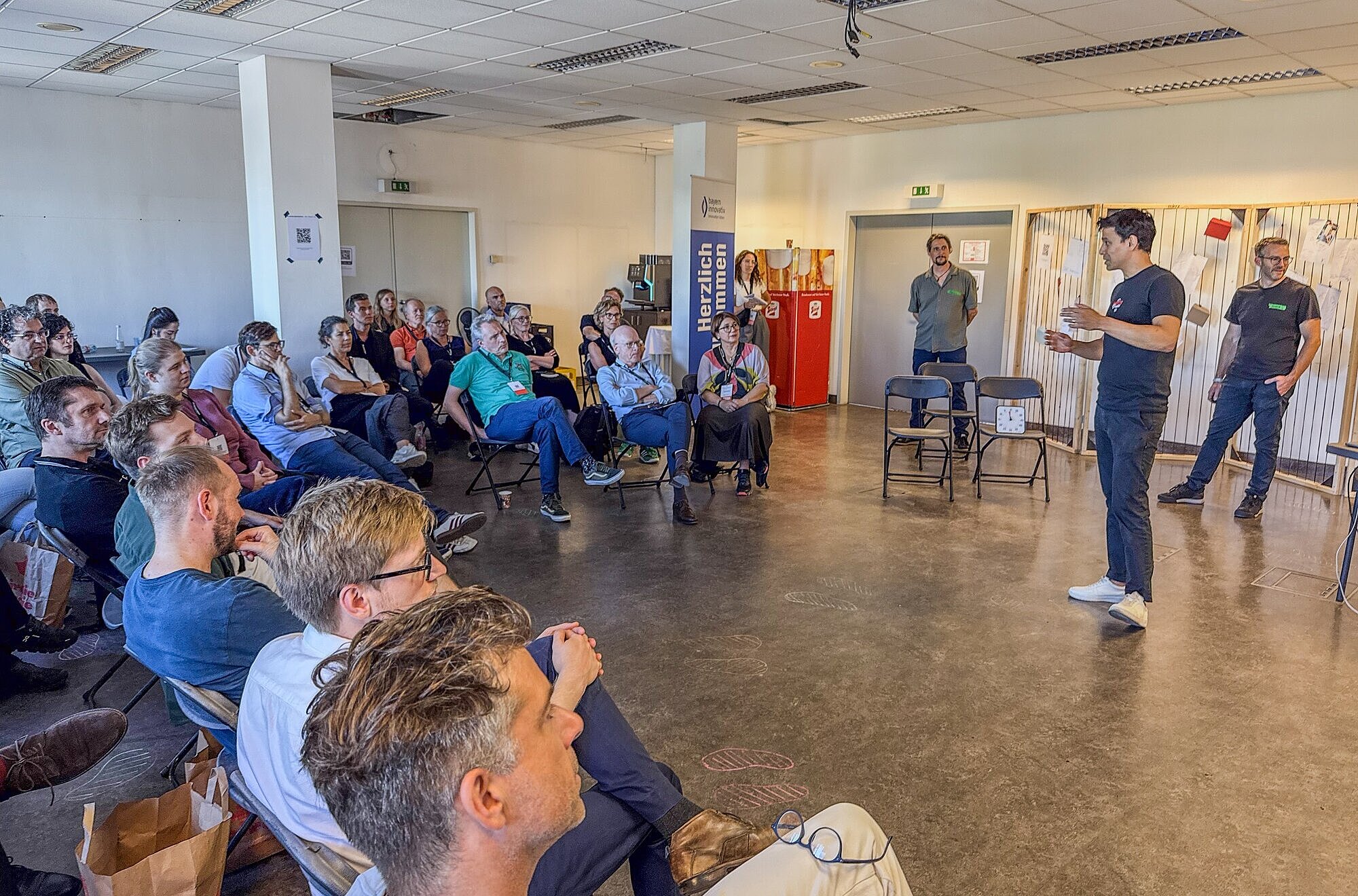 Eine Gruppe von Menschen sitzt in einem Raum und hört einer Person zu, die vorne steht und spricht. Unter den Teilnehmenden sind Johanna Lison, Projekt- und Innovationsmanagerin im Innovationsnetzwerk Kreativwirtschaft der Bayern Innovativ GmbH, Andreas Böhm, Innovationsnetzwerk Digitalisierung, Leitung ThinkNet Quantentechnologie, Bayern Innovativ GmbH und Matthias Konrad, Mitglied der Geschäftsleitung bei der Bayern Innovativ GmbH. Im Hintergrund sind ein Banner mit der Aufschrift „Herzlich willkommen“ sowie weitere Personen zu sehen, die ebenfalls den Ablauf verfolgen.