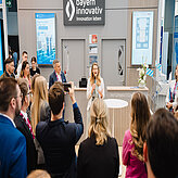 Frau spricht mit Mikrofon vor Publikum bei Messestand mit Schild 'bayern innovativ Innovation leben'