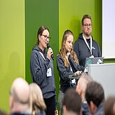 Auf dem Bild sind zwei Frauen und ein Mann zu sehen. Die Frau auf der linken Seite hat ein Mikrofon und hält einen Vortrag.