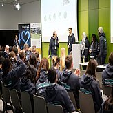 Auf dem Bild ist ein Veranstaltungsraum zu sehen, in welchem einige Personen im Publikum sitzen. Vorne stehen vor einer Leiwand ein Moderator und eine Gruppe von Menschen.