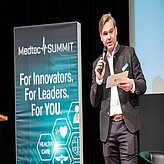 Eine Person steht neben einem Banner und hält ein Mikrofon sowie ein Blatt Papier in der Hand