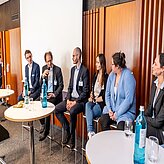 Das Bild zeigt sechs Personen, die nebeneinander auf Stühlen sitzen. Sie sind formell gekleidet und befinden sich offenbar in einem professionellen oder akademischen Umfeld, möglicherweise im Rahmen einer Podiumsdiskussion oder eines Panels.