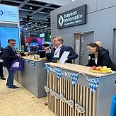 Auf dem Bild sind Personen zu sehen, welcher hinter einer bayrisch geschmückten Theke stehen. Ein Herr im Anzug hat ein Mikrofon in der hat und hält eine Rede.