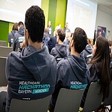 Auf dem Bild ist der Rücken von drei Menschen zu sehen, welche sich einen Vortrag anhören. Sie tragen den gleichen Pullover mit der Aufschrift Healthcare Hackathon Bayern.