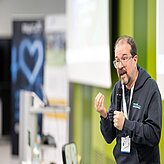 Auf dem Bild ist ein Mann zu sehen, welcher ein Mikro in der Hand hat und einen Vortrag hält. Im Hintergrund sind zwei RollUps und eine Leinwand zu sehen.