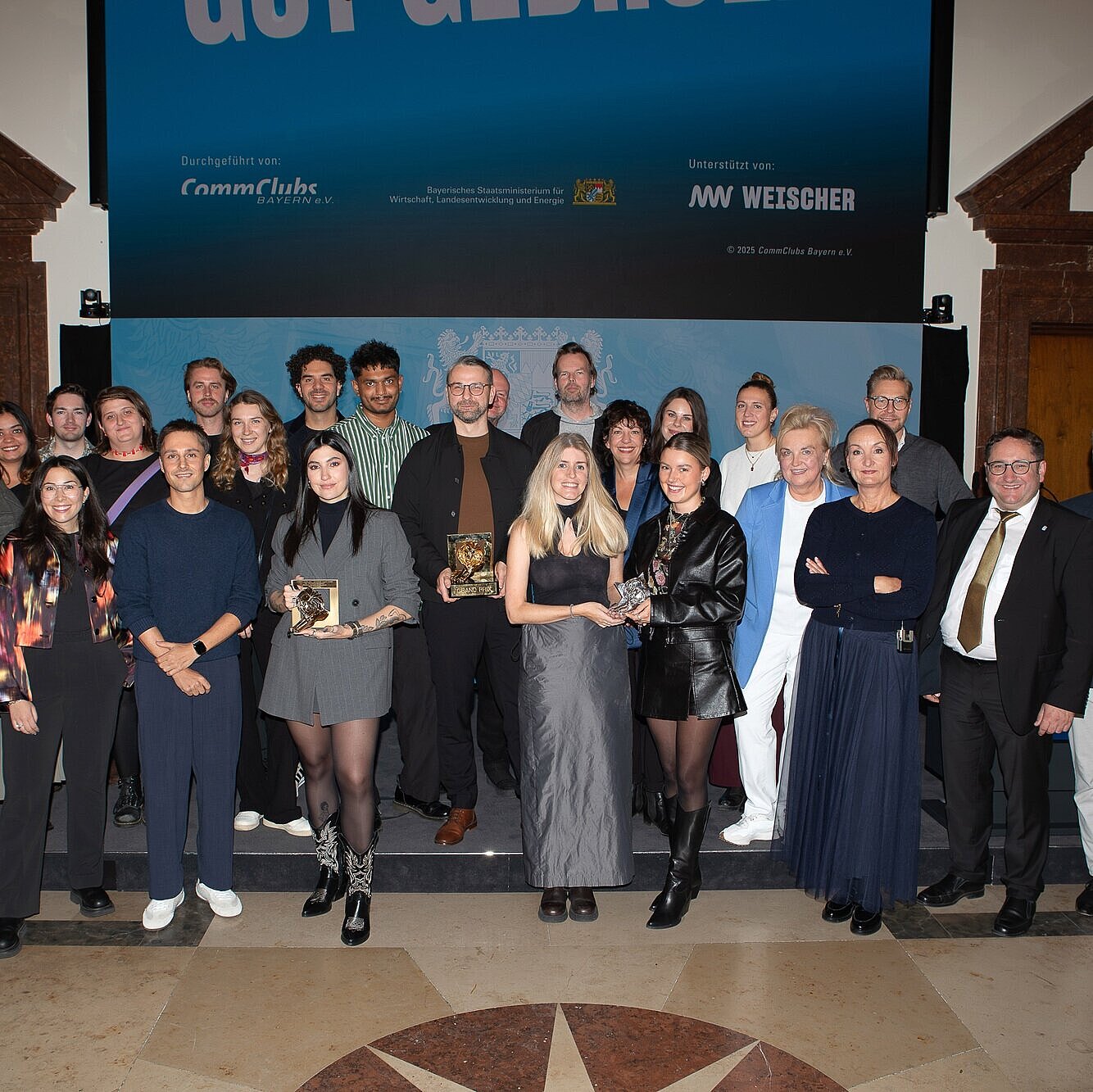 Tobias Gotthardt, Staatssekretär im Bayerischen Staatsministerium für Wirtschaft, Landesentwicklung und Energie, und Petra Lorenz, Geschäftsführerin des CommClubs Bayern e.V., stehen gemeinsam mit bayerischen Gewinnern und Gewinnerinnen des diesjährigen Cannes Lions auf einer Bühne.