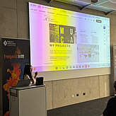 Ánh Nguyen steht neben dem Rednerpult und informiert die Teilnehmenden des Meet-ups über das Thema Curator & Art Consultant. Im Hintergrund sind ein Roll-up und eine Leinwand zu sehen. Die Leinwand wird von mehreren bunten Farben beleuchtet. 