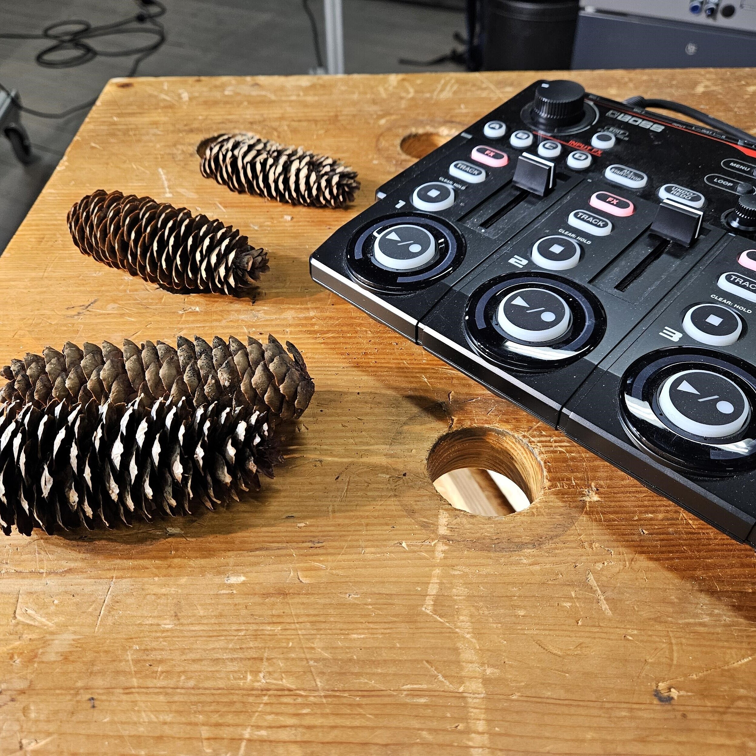 auf einer abgenutzten Holztischplatte liegen vier Tannenzapfen und Ausschnitte eine schwarzen Mischpultes.