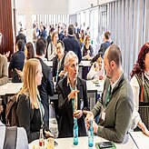 Gruppe von Personen in einem gut beleuchteten Raum, vermutlich im Rahmen einer Konferenz oder Netzwerkveranstaltung. Die Anwesenden sitzen an Tischen und unterhalten sich, einige halten Getränke in der Hand. 