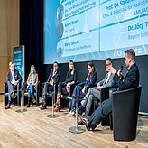 Sieben Personen sitzen auf einer Bühne in Stühlen, vermutlich im Rahmen einer Podiumsdiskussion oder Konferenz. Hinter ihnen befindet sich eine große Leinwand mit Text und Bildern, deren Inhalt jedoch nicht vollständig lesbar ist. Die Teilnehmenden tragen formelle Kleidung