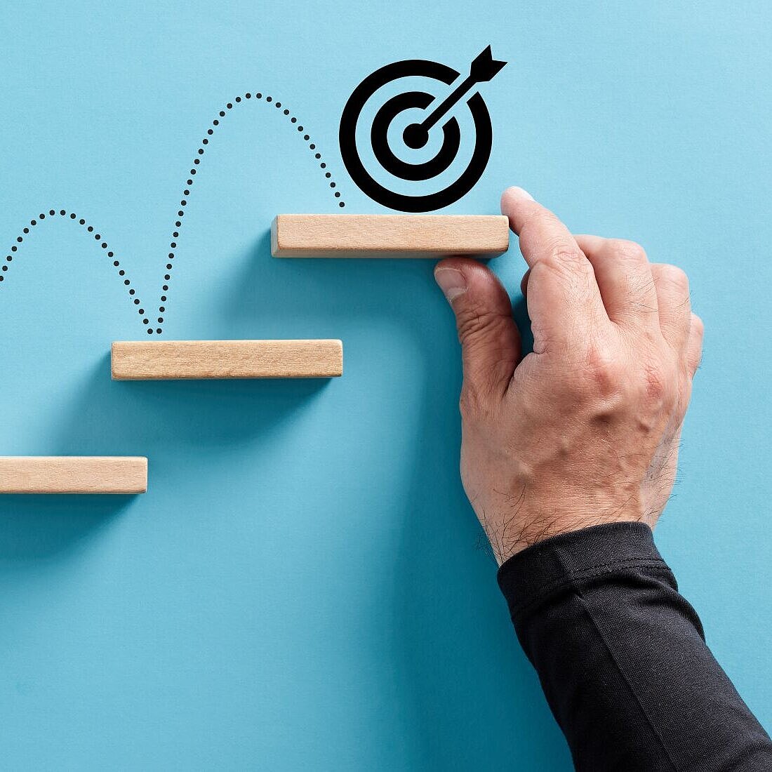 fünf Treppenstufen mit Hüpfandeutungen nach oben, ganz oben hält ist ein Zielsymbol mit einem Kreis und einem Pfeil, eine Hand hält die obereste Stufe