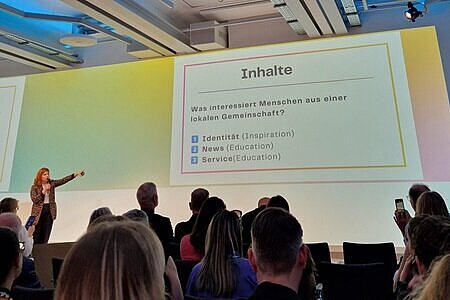 Zu sehen ist die Bühne bei der Transforming Media Veranstaltung, auf der Charline Mohr gerade einen Vortrag hält. Sie blickt und deutet mit ihrer linken Hand in Richtung der Folie, die an die Wand geworfen ist. Das Publikum ist nur in Abrissen von hinten zu erkennen. 