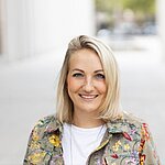 Auf dem Bild ist eine junge Frau mit blonden Haaren zu sehen. Sie trägt ein weißes TShirt und eine bunte Jacke und lächelt freundlich in die Kamera.