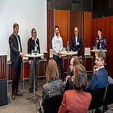 Fünf personen stehen an Stehtischen und unterhalten sich, im Hintergrund eine Leinwand. im Vordergrund Publikum