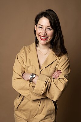 Frau mit schulterlangem Haar trägt beige Jacke, Halskette und Armbanduhr, steht mit verschränkten Armen vor braunem Hintergrund