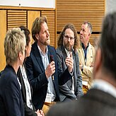 Mehrere Personen sitzen in einer Diskussionsrunde, eine Person hält ein Mikrofon und spricht, im Hintergrund Holzpaneele.