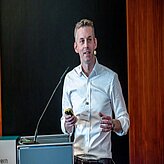 Eine Person steht hinter einem Rednerpult und hält möglicherweise eine Präsentation oder Rede. Die Person hält einige Papiere und eine Fernbedienung in der Hand