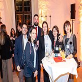 "Eine Gruppe von Personen in formeller Kleidung steht zusammen und posiert für ein Foto bei einer Veranstaltung in Innenräumen. Vor ihnen befindet sich ein Tisch mit Getränken und Snacks. Im Hintergrund sind weitere Gäste, Beleuchtungselemente und Dekorationen zu sehen."