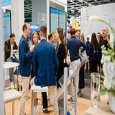 Mehrere Personen stehen und sitzen in einem hellen, modernen Messestandbereich und unterhalten sich