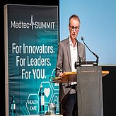 das Bild zeigt eine Person, die an einem Rednerpult steht und in ein Mikrofon spricht. 