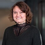 Auf dem Bild ist eine Frau mit braunen, schulterlangen Haaren zu sehen. Sie trägt ein schwarzes, hochgeschlossenes Oberteil und lächelt freundlich in die Kamera.