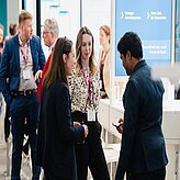 Gruppe von Menschen mit Namensschildern im Gespräch vor einem Bildschirm mit Text in einem hellen Innenraum