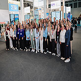 Große Gruppe von Menschen unterschiedlichen Alters und Geschlechts in einem Messe- oder Ausstellungsraum mit Ständen im Hintergrund