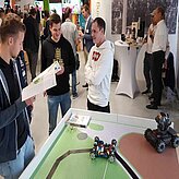 In der Ausstellung stehen an einem Stand 3 Männer im Gespräch vertieft