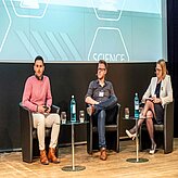 Drei Personen sitzen auf einer Bühne jeweils in einem schwarzen Sessel. Vor ihnen stehen kleine Tische mit Wasserflaschen. I