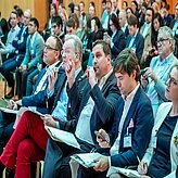Bild zeigt eine Gruppe von Personen, die in einem Konferenz- oder Seminarumfeld sitzen. Die Anwesenden tragen Businesskleidung, einige halten Notizblöcke oder Unterlagen in der Hand.