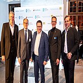 Fünf Personen in formeller Kleidung stehen vor einem Hintergrund mit verschiedenen Logos.