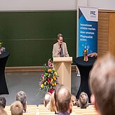 Person steht hinter einem Rednerpult vor einem grünen Tafel-Hintergrund und einem Banner mit dem Schriftzug 'PPZ NÜRNBERG Innovationen erlebbar machen Ideen umsetzen Pflegequalität sichern'