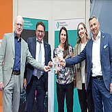 Fünf Personen stehen nebeneinander und blicken in Richtung Kamera. Sie tragen formelle bis halbformelle Kleidung und halten ein Hashtag Zeichen in der Hand