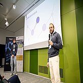 Auf dem Bild sind zwei Männer zu sehen, welcher vor einer Leinwand stehen und einen Vortrag halten.