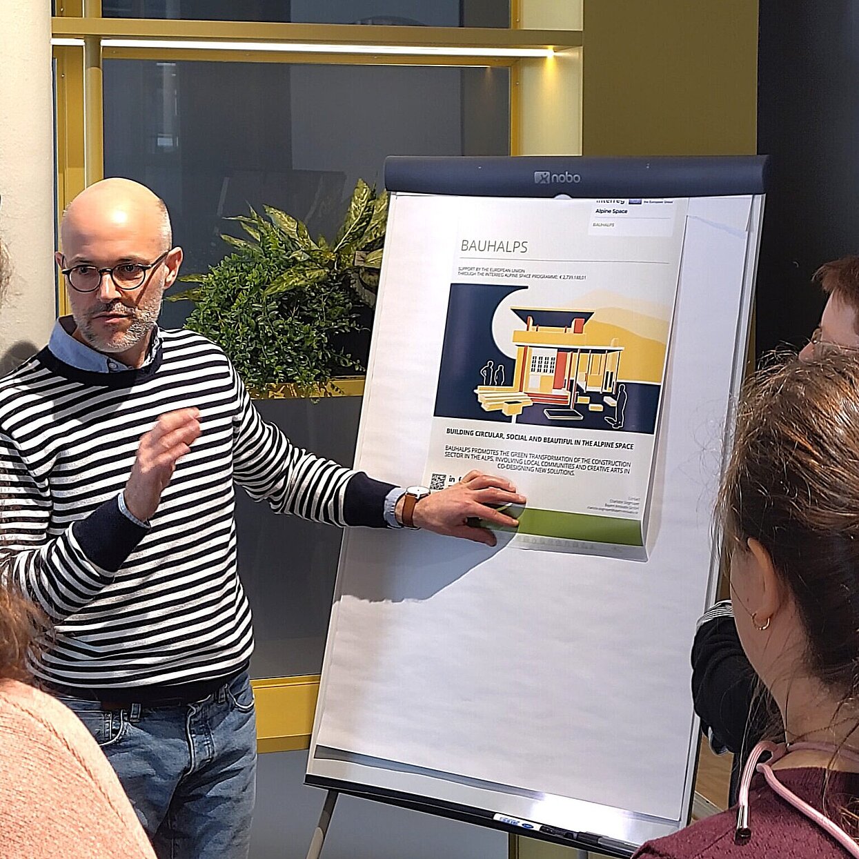 Ein Mann steht vor einem Plakat vor auf einem Schreibboard und erklärt etwas. 